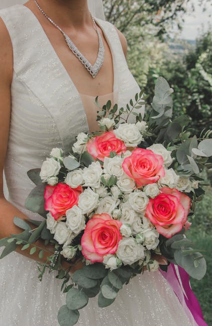 Close-up of a bride wearing a white, sleeveless wedding dress with subtle shimmer, holding a large floral bouquet. The bouquet features vibrant pink roses, white roses, and small white filler flowers,